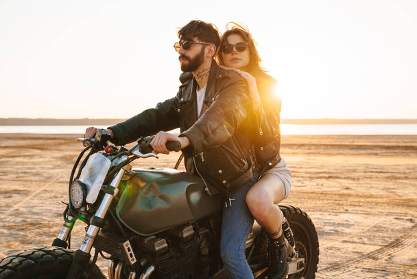 Beautiful young stylish couple enjoying ride on a motorbike at the beach - 写真・画像