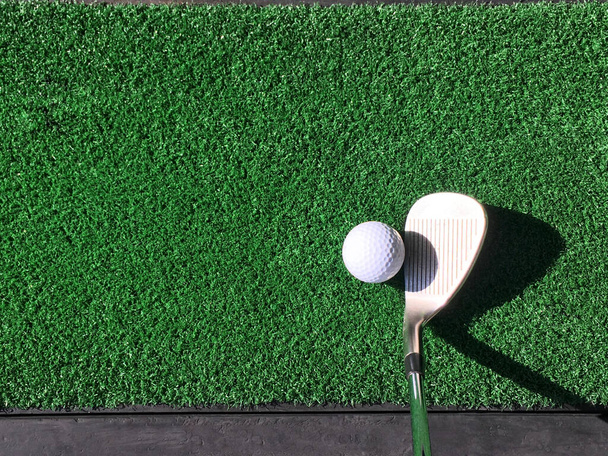 Ein weißer Golfball mit keileisernem Schläger auf Übungs-Kunstrasenmatte unter Morgensonne - Foto, Bild