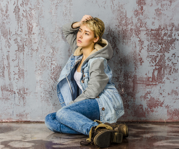 Young beautiful girl with short hair in jeans and a denim jacket sitting at the wooden wall - Foto, afbeelding