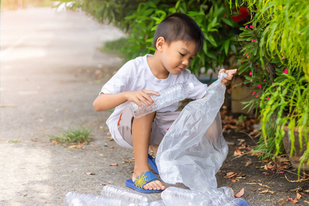 Στο δημόσιο πάρκο της πόλης. Το παιδί από την Ασία είναι εθελοντής για να καθαρίσει το πάτωμα. Μαζεύει πολλά πλαστικά μπουκάλια και άχυρα στο έδαφος. Αποθήκευση περιβαλλοντικών και μείωση της έννοιας των αποβλήτων. - Φωτογραφία, εικόνα