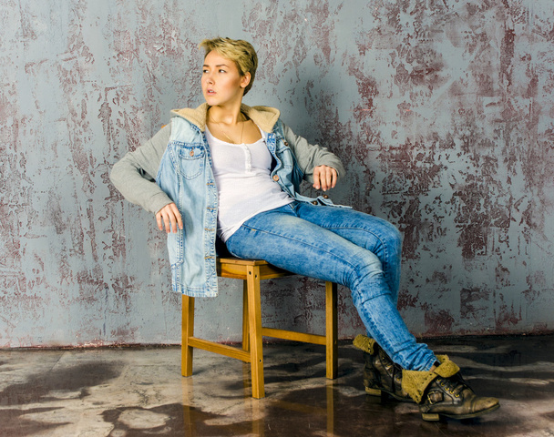 Young beautiful girl with short hair in jeans and a denim jacket sitting at the wooden wall - Fotografie, Obrázek