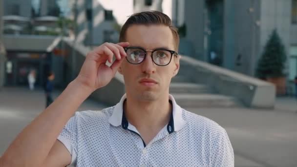 Σοκαρισμένος νεαρός επιχειρηματίας βγάζει τα γυαλιά του αιφνιδιαστικά. Έκπληκτος άνθρωπος που φοβάται από τις ειδήσεις κοιτάζει την κάμερα στο παρασκήνιο της επιχειρηματικής περιοχής της πόλης - Πλάνα, βίντεο