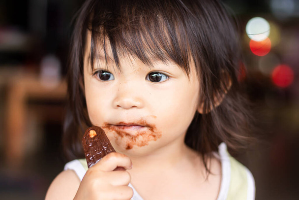 Portrét 1 až 2 let starého dítěte. Šťastné asijské dítě dívka jíst zmrzlinu hnědou čokoládu sama. Její tvář špinavá a špinavá jídlem. - Fotografie, Obrázek