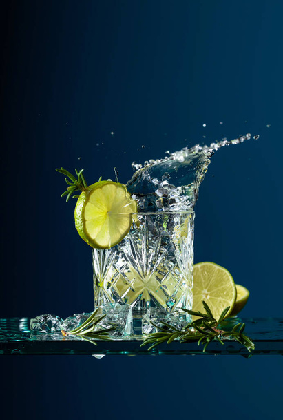 Gin-tónico com limão e alecrim. Bebida carbonatada com pedaços de gelo em uma mesa de vidro. Fundo azul, espaço de cópia
. - Foto, Imagem