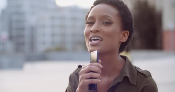 Portrait of happy American woman singing song in phone as in microphone. African ethnic lady in casual short haired dress smiling dancing to music in street, one person party, urban scene, close up - Footage, Video