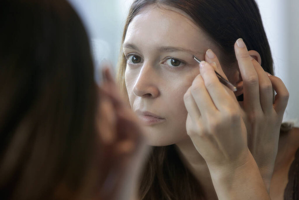 Junge Kaukasierin zupft sich vor dem Spiegel die Augenbrauen. - Foto, Bild