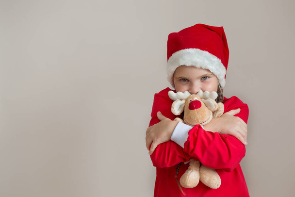 Açık gri arka planda geyik oyuncağı olan Noel Baba kostümlü mutlu kız.  - Fotoğraf, Görsel