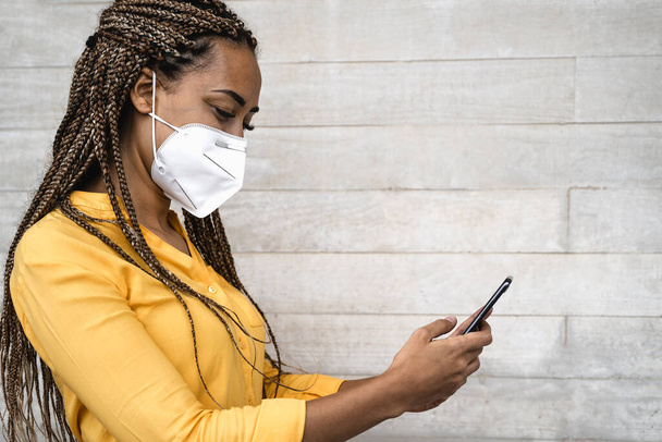 Africká žena na obličeji lékařské masky pomocí mobilního smartphonu - Mladá dívka s copánky baví s telefonem během propuknutí viru corona - Zdravotní lidé a koncept technologie - Fotografie, Obrázek
