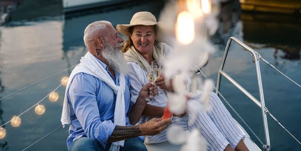 Senior couple toasting champagne and eating fruits on sailboat vacation - Χαρούμενοι ηλικιωμένοι που διασκεδάζουν γιορτάζοντας την επέτειο του γάμου τους στο ταξίδι - Αγάπη και ταξιδιωτικός τρόπος ζωής - Φωτογραφία, εικόνα