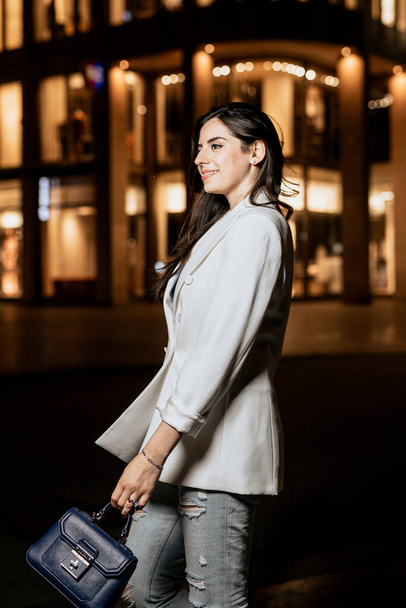 Foto vertical de una mujer atractiva con chaqueta de moda blanca y jeans mirar hacia otro lado. Vista lateral de la mujer joven en el edificio con fondo de iluminación - Foto, Imagen