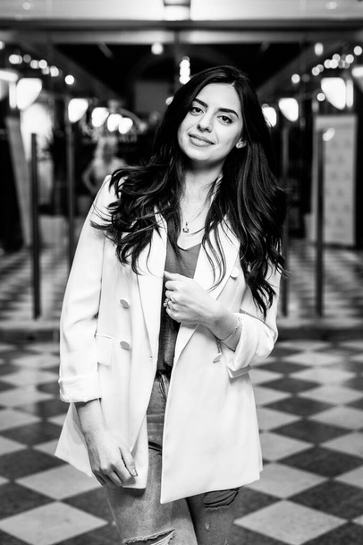 Mulher sorrindo atraente vestindo jaqueta de moda branca e jeans olhar na câmera. preto e branco foto de jovem mulher no moderno loja de fundo - Foto, Imagem