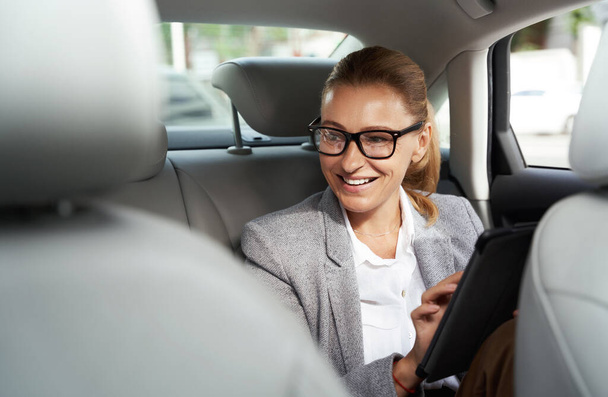 Ofise giden yolda çalışıyorum. Başarılı bir iş kadını dijital tablet kullanarak gözlük takıyor ve arabada arka koltukta otururken gülümsüyor. - Fotoğraf, Görsel