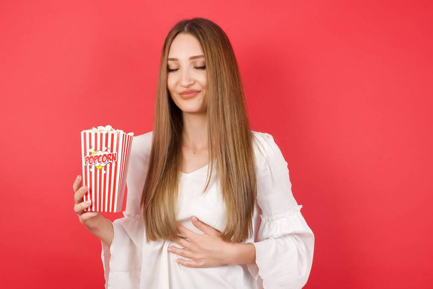 Giovane donna caucasica che tiene secchio con popcorn in piedi sopra isolato sfondo rosso tocca pancia, sorride dolcemente, mangiare e concetto di soddisfazione. - Foto, immagini