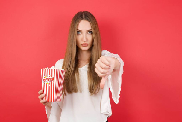 Young beautiful woman with popcorn being upset holding thumb down. Dislike concept.  - Фото, изображение