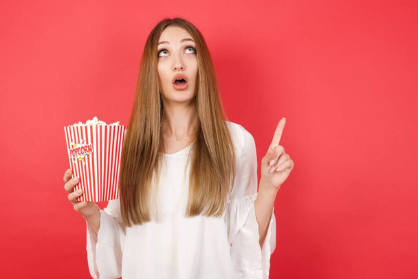 Young beautiful woman with popcorn surprised looking up and pointing with fingers and raised arms. - Foto, imagen