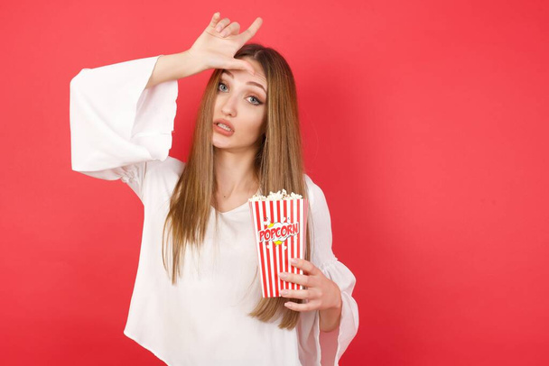 Young beautiful woman with popcorn making fun of people with fingers on forehead doing loser gesture mocking and insulting. - Photo, Image
