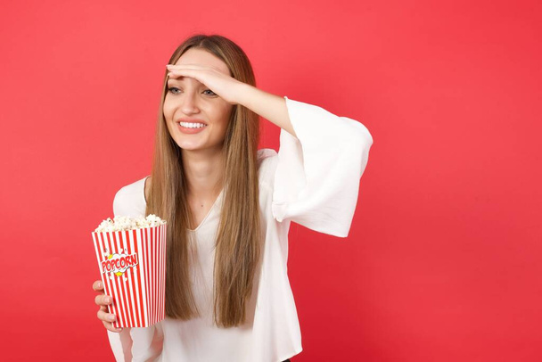 Young beautiful woman with popcorn very happy and smiling looking far away with hand over head. Searching concept. - Foto, Imagem