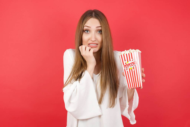 Jonge mooie vrouw met popcorn houdt handen dicht bij de mond, voelt zich bang en bang, heeft een fobie, shock en angstig concept. - Foto, afbeelding
