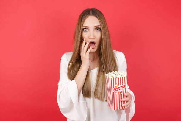 Studio shot van bange jonge mooie vrouw met popcorn. Het begrip mens en menselijke emoties - Foto, afbeelding