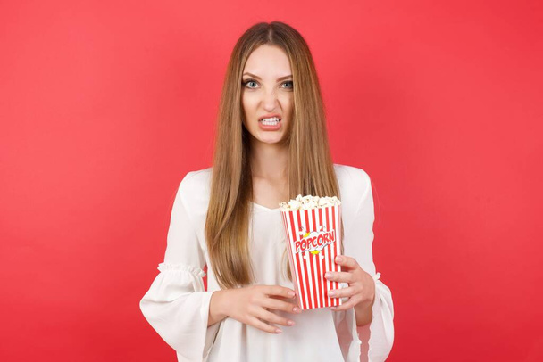 jonge mooie vrouw met popcorn houdt tanden gebald, fronst gezicht in ontevredenheid  - Foto, afbeelding
