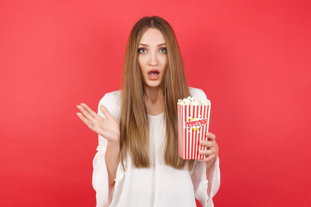 Surprised terrified young beautiful woman with popcorn gestures with uncertainty, stares at camera, puzzled as doesn't know answer on tricky question, People, body language, emotions concept - Photo, image