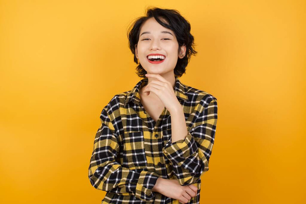 Optymistyczny uśmiechnięty szczęśliwy azjatycki kobieta w żółty sweter pozowanie w studio na żółtym tle  - Zdjęcie, obraz
