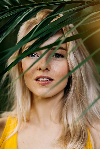 Colorful closeup photo of young beautiful blonde model in yellow swimsuit smiling posing standing under evergreen tree on sunny summer day. Cosmetics, beauty, skin care, vacation concept. - Foto, Imagem