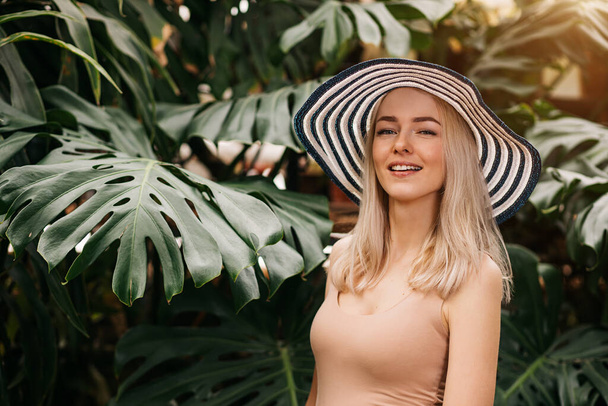 Lovely amazed blonde lady wearing sun hat, swimsuit, he smiles and looks at the camera from copy space, standing on tropical plants background. Summer vacation, facial expressions, emotion concept. - Photo, Image