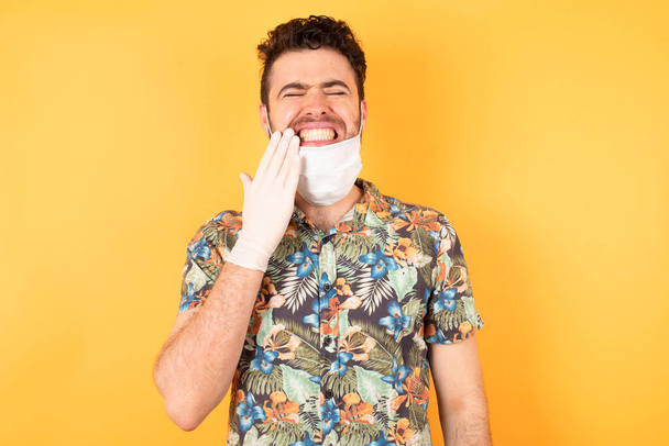 Mladý pohledný muž v ochranné masce dotýkající se úst s rukou s bolestivým výrazem, protože bolest zubů nebo zubní onemocnění na zubech. - Fotografie, Obrázek