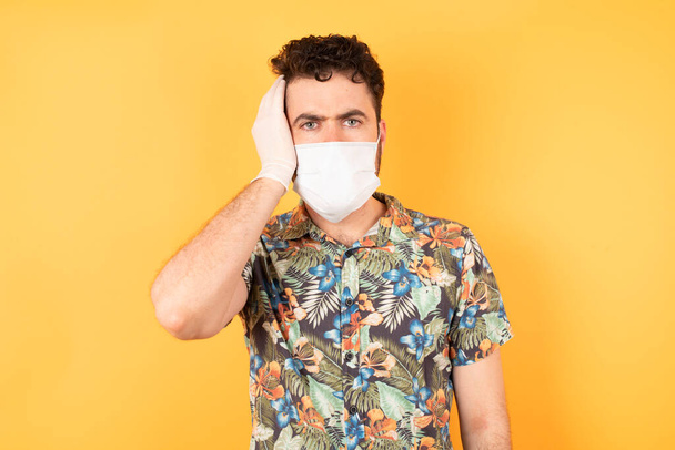 Caucasian man in medical mask in yellow studio, shocked look, hand on face  - Photo, Image