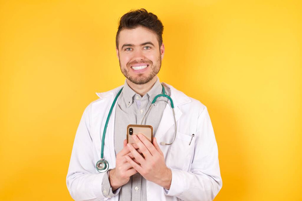 Zdjęcie lekarza człowiek cieszy się odległą komunikacją, używa telefonu komórkowego, surfuje szybki nieograniczony internet, ma przyjemny uśmiech, noszenie munduru medycznego, sprawia, że zakupy online - Zdjęcie, obraz