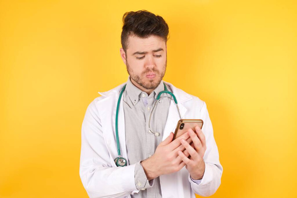 Portret zaimponowanego kaukaskiego lekarza używa telefonu komórkowego, czyta niesamowite wiadomości w mediach społecznościowych, krzyczy wow omg, żółte tło - Zdjęcie, obraz