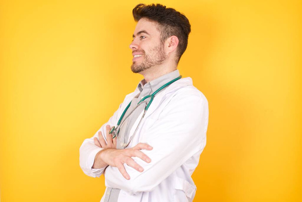 Porträt eines fröhlichen kaukasischen jungen Arztes, der die Arme verschränkt und beiseite schaut, während er ein nettes Gespräch mit seinem Patienten führt. Tragt Uniform und Stethoskop isoliert über gelbem Hintergrund - Foto, Bild