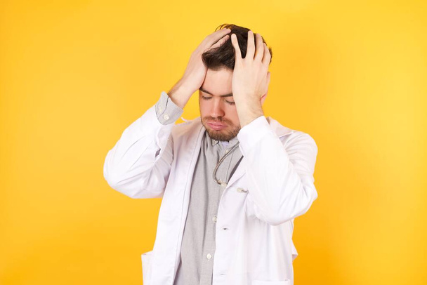 Fiatal jóképű fehér orvos férfi orvosi egyenruhát visel szenved erős fejfájás kétségbeesett és stresszes, mert a túlzott munka. Depresszió és fájdalom koncepció. - Fotó, kép