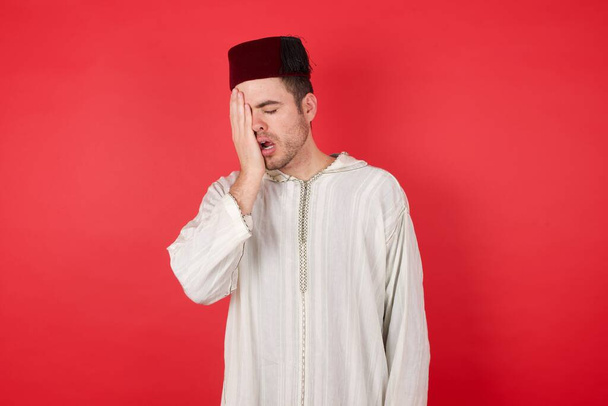 schöner junger Mann mit traditionellem türkischen Hut Fez gähnend müde, das halbe Gesicht, Augen und Mund mit der Hand bedeckt. Gesicht schmerzt vor Schmerzen. - Foto, Bild