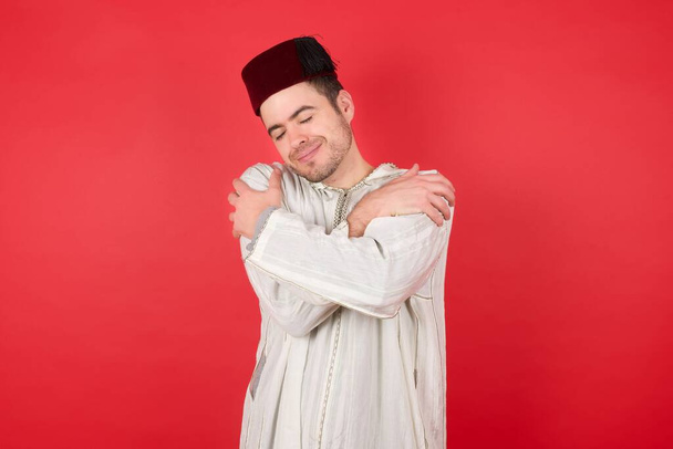 όμορφος νεαρός άνδρας που φοράει παραδοσιακό τούρκικο καπέλο φέσι αγκαλιάζοντας τον εαυτό του χαρούμενο και θετικό, χαμογελώντας με αυτοπεποίθηση. Αυτοαγάπη και φροντίδα - Φωτογραφία, εικόνα
