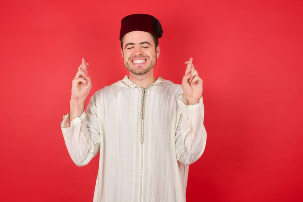 joven guapo con el tradicional sombrero turco fez gesto cruzado, sonriendo con esperanza y los ojos cerrados. Suerte y concepto supersticioso. - Foto, imagen