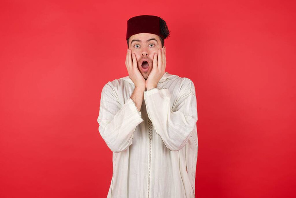 όμορφος νεαρός άνδρας φορώντας παραδοσιακά τούρκικα καπέλο φέσι εκφράζει ενθουσιασμό και συγκίνηση, κρατά το σαγόνι έπεσε, τα χέρια στα μάγουλα, έχει τα μάτια πεταμένα έξω, φοράει κόκκινο πουκάμισο, μοντέλα εσωτερική μόνο - Φωτογραφία, εικόνα