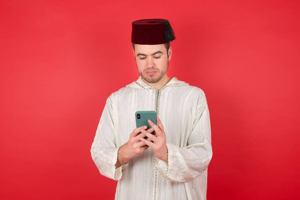 handsome young man wearing traditional turkish hat fez using smartphone over red background - Valokuva, kuva