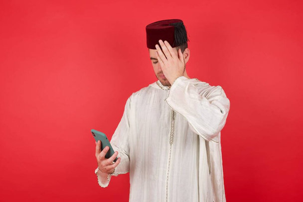 hombre musulmán triste en el estudio sosteniendo el teléfono móvil, mano a cara  - Foto, imagen