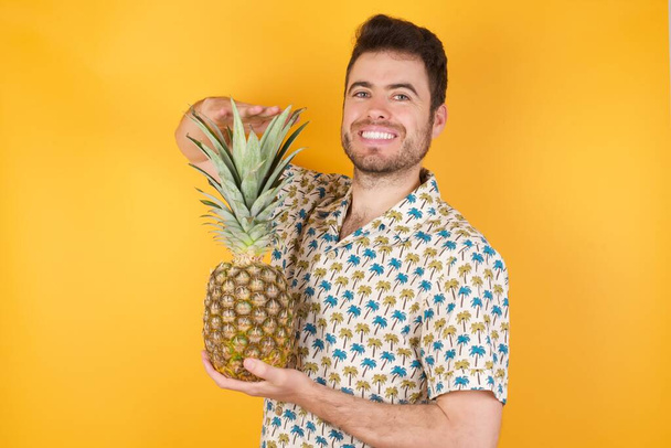Handsome young man posing with pineapple over yellow background. Studio shot - Photo, image