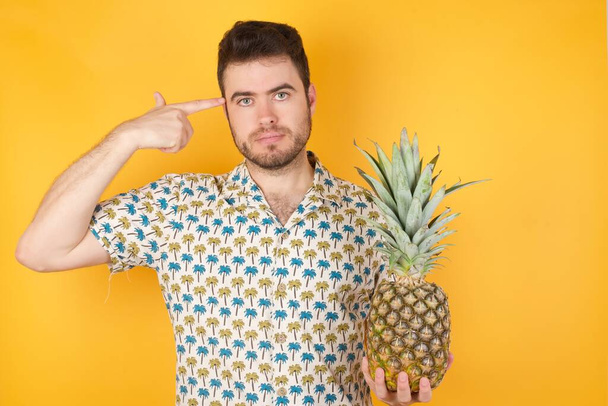 Handsome young man posing with pineapple over yellow background. Studio shot - Foto, immagini