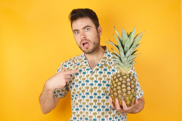 jonge man met ananas geeft op zichzelf met verbijsterde uitdrukking, wordt geschokt   - Foto, afbeelding