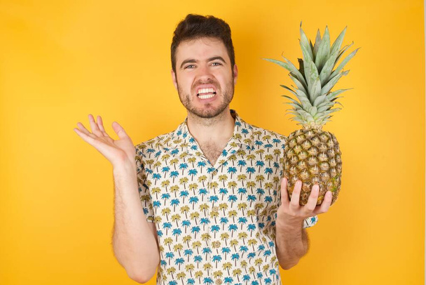 молодий чоловік з ананасовими жестами в здивуванні, заморожений обличчям з невдоволенням. Негативні емоції та вираз обличчя
 - Фото, зображення