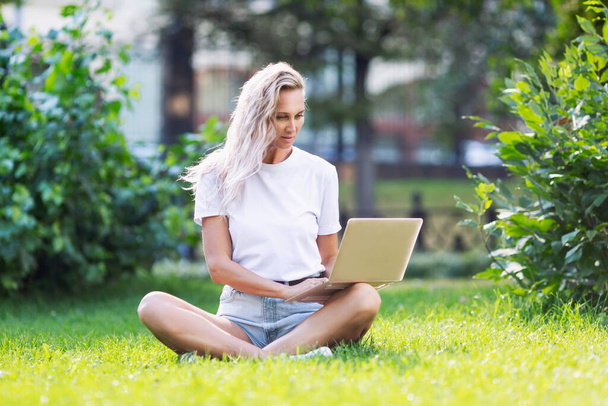 Junge Frau mit Laptop sitzt auf dem Rasen in einem Sommerpark. Blonde mit langen Haaren in weißem Tank-Top und kurzer Hose. Sonniger Tag. Fernarbeit und Fernstudium. - Foto, Bild