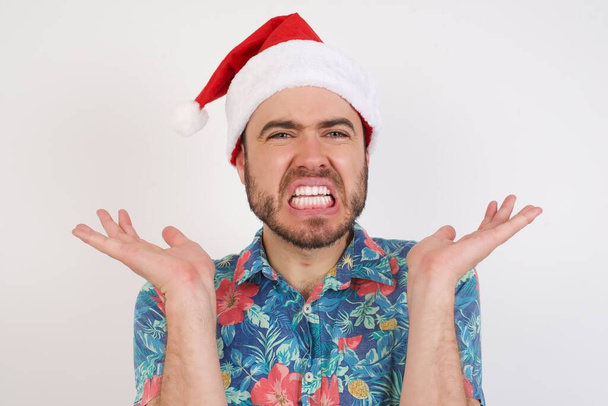 Jonge man in Santa hoed gebaren in verbijstering, fronst gezicht met ontevredenheid. Negatieve emoties en gezichtsuitdrukking - Foto, afbeelding