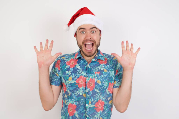 Noel Baba şapkalı genç adam sevinçten avuçlarını kaldırıyor, birinden müthiş bir hediye almaktan mutlu, yüksek sesle bağırıyor, heyecanlı beyaz erkek çığlıkları atıyor.. - Fotoğraf, Görsel