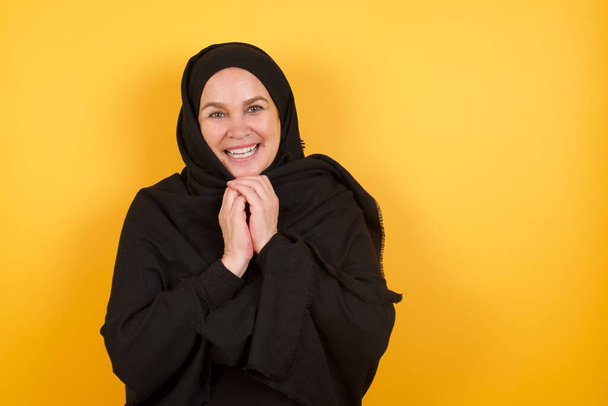 Зрелая женщина, носящая хиджаб, радостно улыбается, радуется приятным новостям от собеседницы, держит руку на пульсе. Эмоции людей. - Фото, изображение