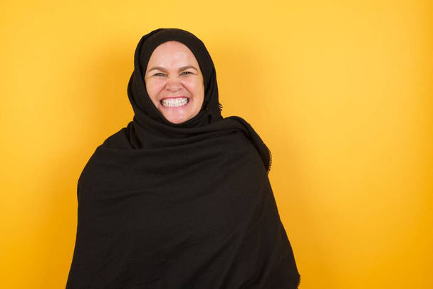 Volwassen vrouw draagt hijab houdt tanden gebald, fronst gezicht in ontevredenheid, geïrriteerd vanwege veel taken. Woedende man kijkt uit het raam, ziet ellendig weer - Foto, afbeelding