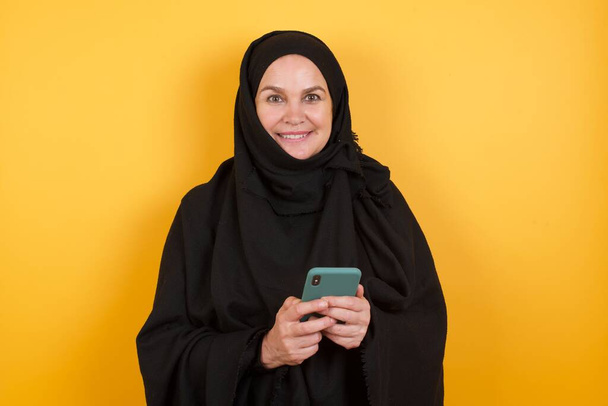 Зріла жінка в хіджабі використовує мобільний телефон, сервери швидкий безмежний Інтернет, має приємну посмішку, одягнену в червону сорочку, робить покупки в Інтернеті, ізольовані на жовтому
 - Фото, зображення